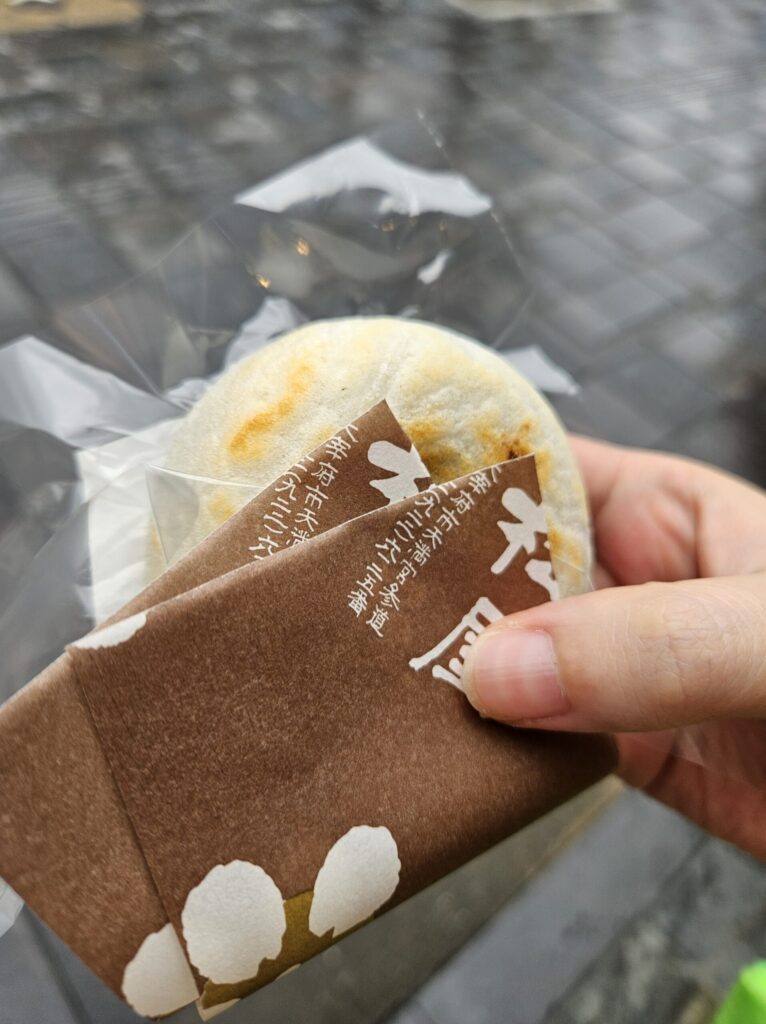 Close-up of a freshly grilled Umegae Mochi from Matsuya held in the right hand with its brown paper wrapper — the golden surface peeking through the wrapper captures the warmth and charm of Dazaifu’s famous sweet.