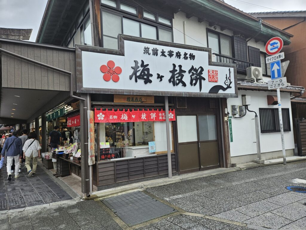 Exterior of Matsuo Shoten, a long-established Umegae Mochi shop along the approach to Dazaifu Tenmangu Shrine. The wooden signboard and nostalgic storefront reflect a warm, handmade charm of traditional Dazaifu.