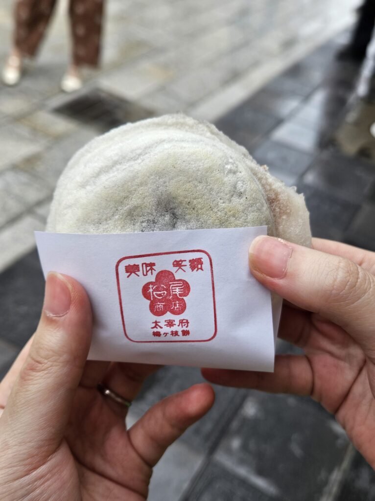 Close-up of a handmade Umegae Mochi from Matsuo Shoten, gently held in both hands with its white paper wrapper — the simple, nostalgic look captures the warmth and care of Dazaifu’s traditional sweet.