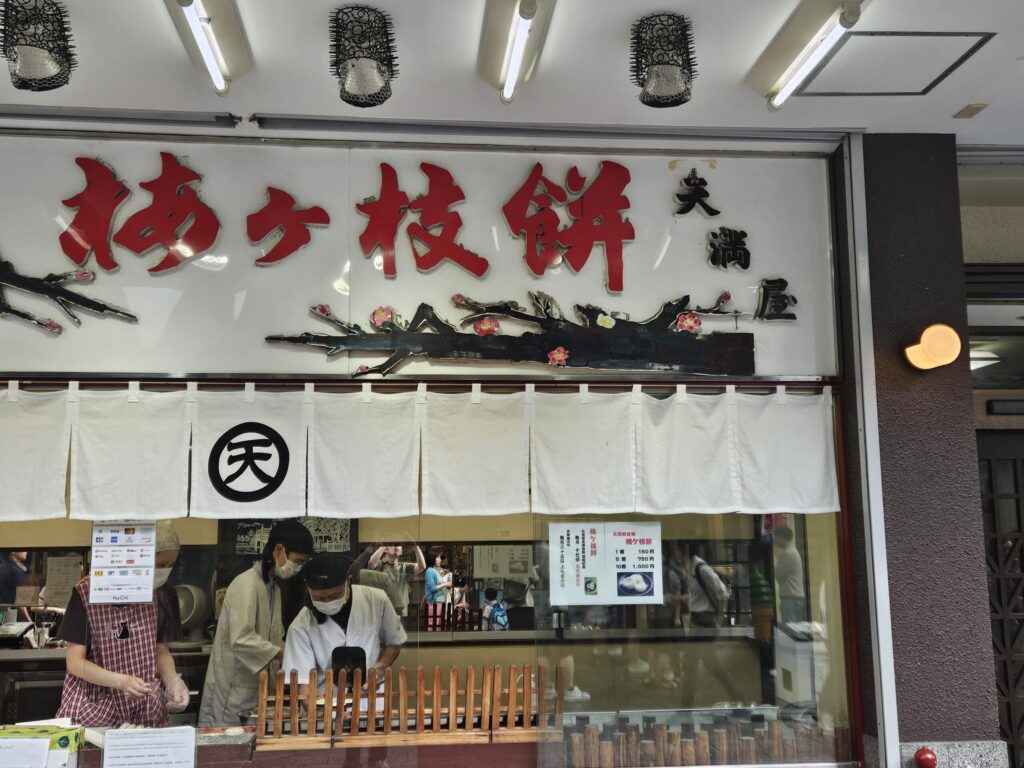 Exterior of Tenmaya, a long-established Umegae Mochi shop on the approach to Dazaifu Tenmangu Shrine. The air is filled with the toasty aroma of freshly grilled mochi as visitors stop by to enjoy the shop’s famous Umegae Mochi right off the grill.