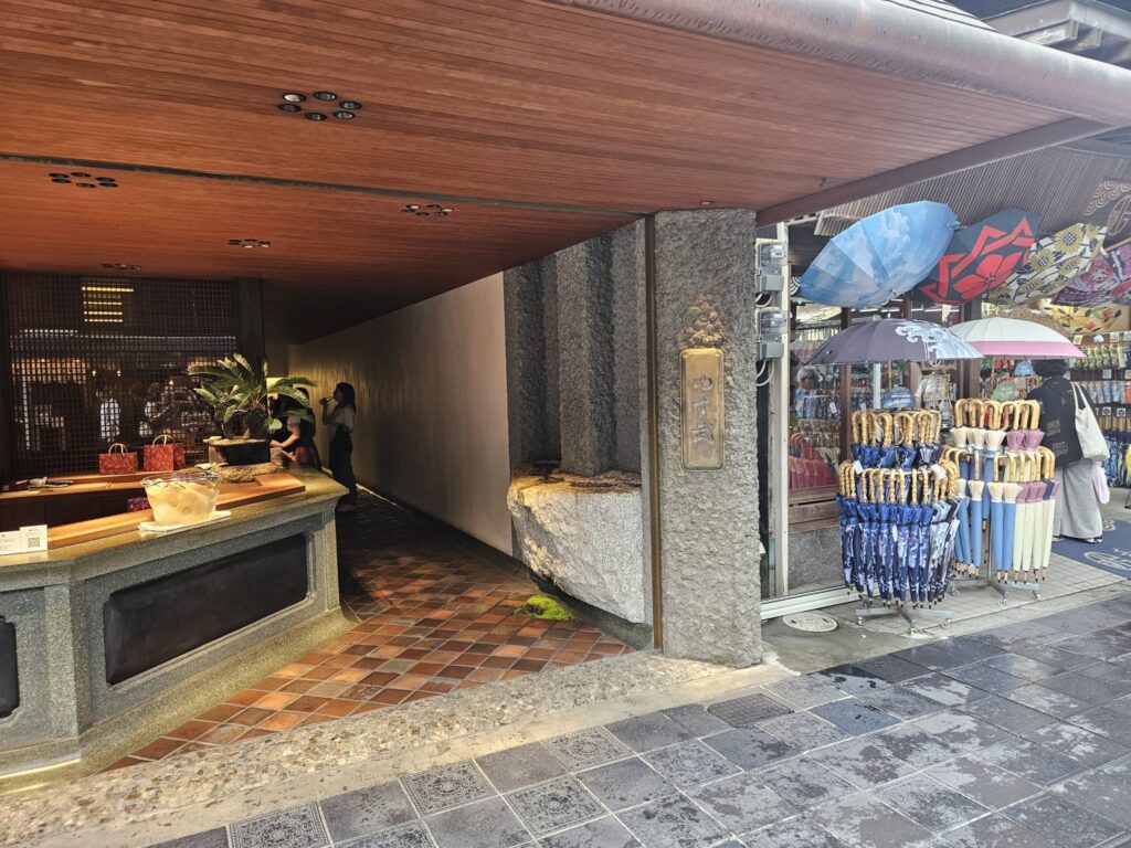 Exterior of Yasuke, a refined Umegae Mochi shop on the approach to Dazaifu Tenmangu Shrine. The storefront features an elegant, clean design with displays of plum cider and souvenirs, attracting visitors with its calm and sophisticated atmosphere.