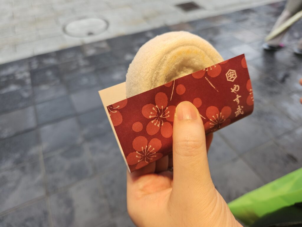 Close-up of a Umegae Mochi from Yasuke, held in the left hand with its elegant washi-style wrapping. The soft pastel tones and refined design reflect the shop’s calm, sophisticated charm unique to this traditional Dazaifu sweet.