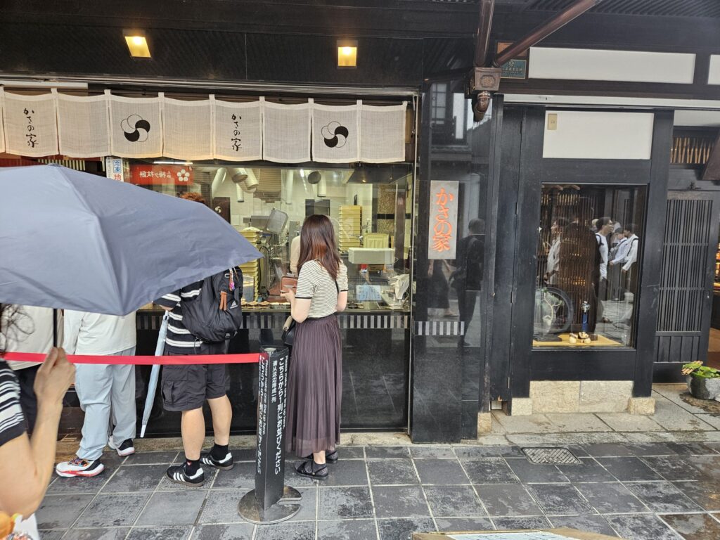 Exterior of Kasanoya, a famous long-established Umegae Mochi shop on the approach to Dazaifu Tenmangu Shrine. Visitors line up in front of the shop for freshly grilled mochi, filling the air with a warm, toasty aroma.