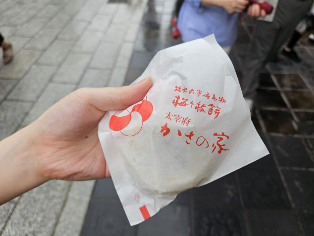 Photo of a Umegae Mochi from Kasanoya held in the right hand, wrapped completely in white paper — the mochi itself isn’t visible, but it captures the joy of finally getting one from this famous, always-busy Dazaifu shop.