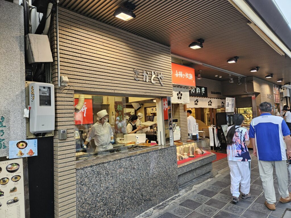 Exterior of Sakadoya, a lively Umegae Mochi shop and restaurant on the approach to Dazaifu Tenmangu Shrine. In addition to mochi, it serves rice bowls, soba, and plum soft-serve ice cream, attracting many visitors throughout the day.