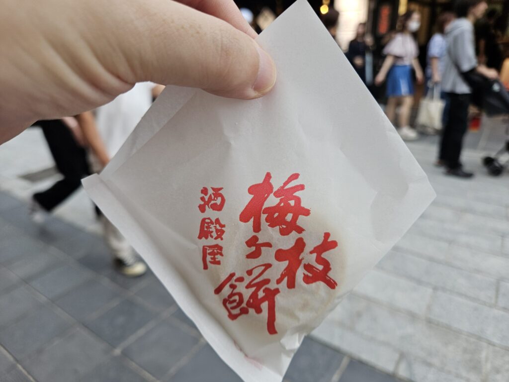 Photo of a freshly grilled Umegae Mochi from Sakadoya, held from the top with its white paper wrapper — the mochi itself isn’t visible because it’s piping hot, but the image captures the feeling of holding a just-made treat from this popular Dazaifu shop.
