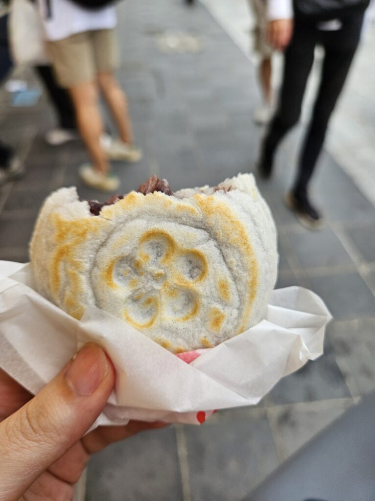 Close-up of a Umegae Mochi from Sakadoya, partially bitten to reveal the sweet red bean filling. The plum-shaped branding mark on the surface is clearly visible, showing the handmade texture and freshly grilled aroma of this Dazaifu specialty.