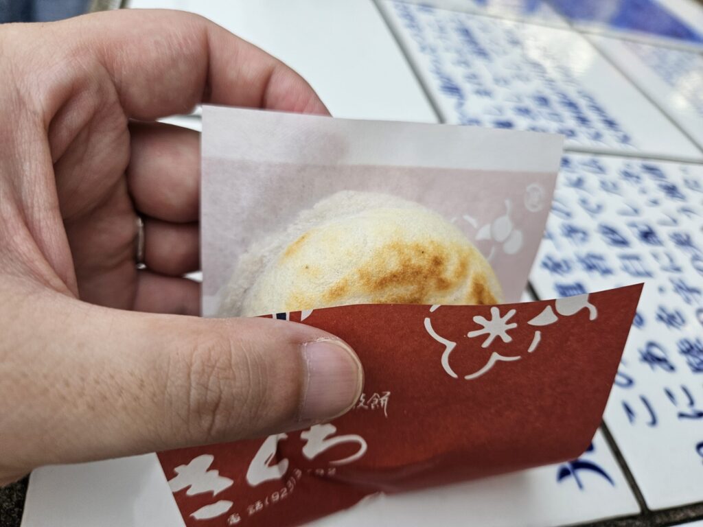 Close-up of a Umegae Mochi from Kikuchi, held in the left hand with its brown paper wrapper. The golden surface peeking through the wrapper captures the freshly grilled warmth and inviting aroma of this popular Dazaifu treat.