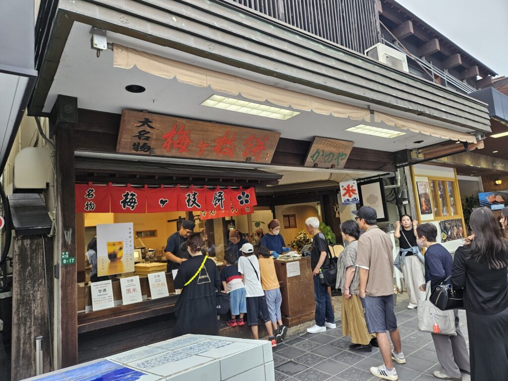 Exterior of Kanoya, a cheerful Umegae Mochi shop on the approach to Dazaifu Tenmangu Shrine. Visitors can watch the staff hand-grill mochi at the storefront, and the shop is also known for its refreshing “Dazaifu Squash,” a fizzy plum drink popular among tourists.