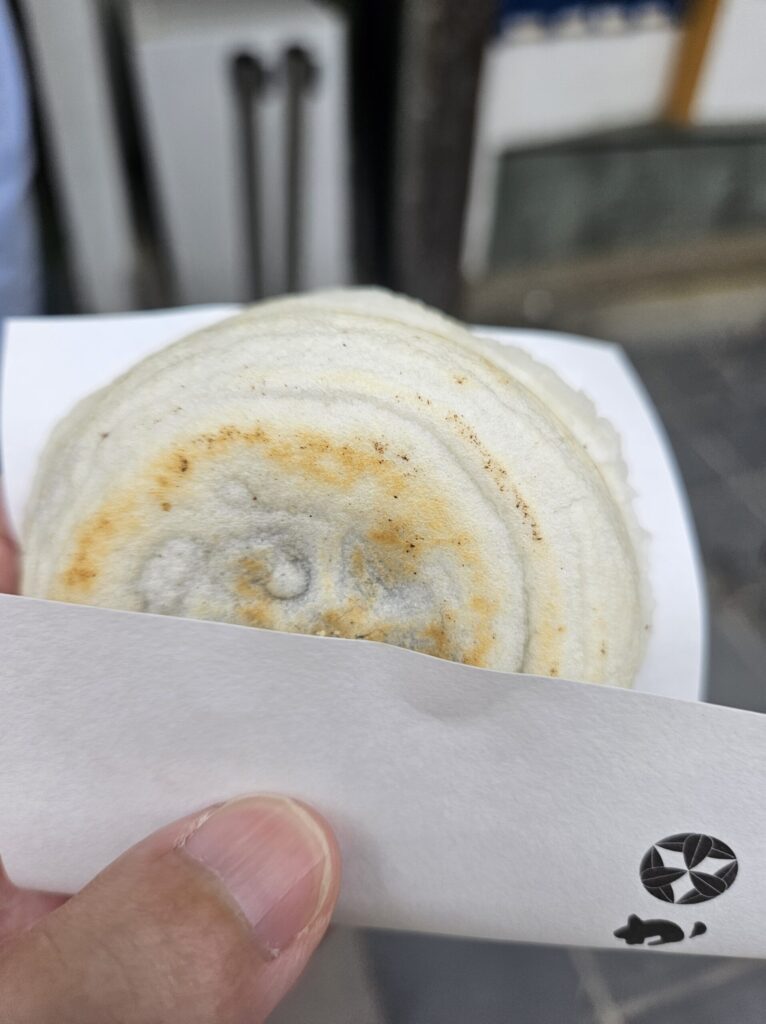 Close-up of a freshly hand-grilled Umegae Mochi from Kanoya, held in the left hand with its white paper wrapper. The warmth and toasty aroma from the freshly made mochi can almost be felt through the wrapper.