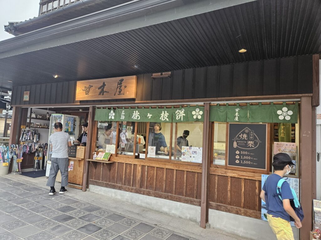 Exterior of Amagiya, a traditional Umegae Mochi shop on the approach to Dazaifu Tenmangu Shrine. The air around the storefront is filled with the cozy aroma of roasted chestnuts and freshly grilled mochi, creating a warm, nostalgic atmosphere.