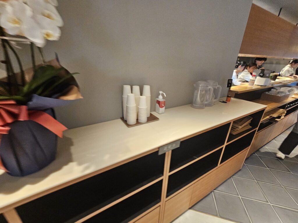 Self-service water station with paper cups and a water pitcher inside nana’s green tea Dazaifu Sando Café.