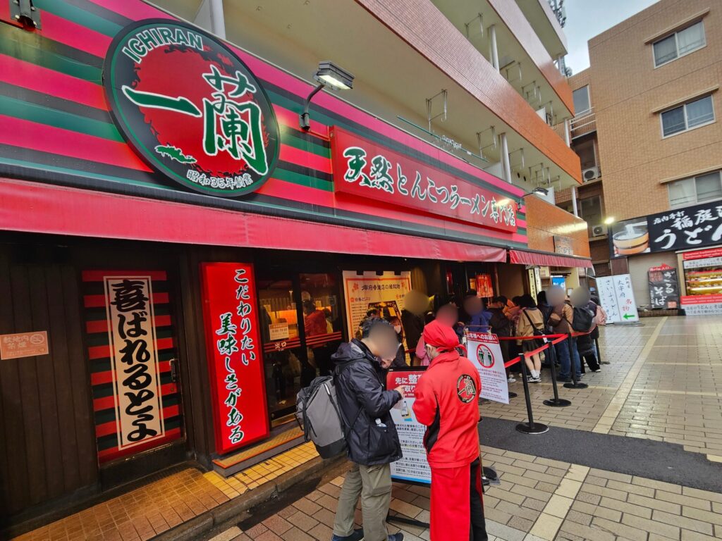 Ichiran Dazaifu Sando Ten exterior with its iconic red sign, customers lining up outside, and a staff member giving explanations near the entrance.